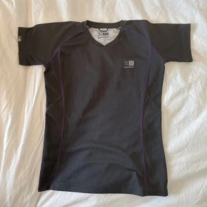 Svart tränings-tshirt från Karrimor - Sportig svart t-shirt från Karrimor med lila sömmar och diskret logga på bröstet. Modellen är kortärmad med rund hals och tillverkad i ett lätt syntetmaterial som andas, perfekt för träning. Snygg passform som sitter bra på kroppen.