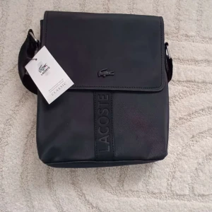 Svart axelväska från Lacoste - Säljer min Lacoste väska eftersom den skulle aldrig passa mig den ser lite fånig ut på mig.   Färgen är svart och möts bara i Märsta!