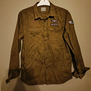 Olivgrön skjorta - Snygg olivgrön skjorta från Primark med feta patchar på bröstet och ärmen, bland annat 'Pacific' och 'Brooklyn'. Skjortan har två bröstfickor med knappar och klassisk krage. Perfekt för som både är intresserad av militär stil och samt grish!