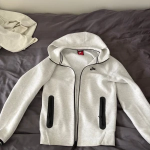 Nike tech fleece - Nike hoodiejacka i vit med svarta detaljer och dragkedjor på fickorna. Jackan har huva, lång ärm och svart Nike-logga på bröstet. Materialet är mjukt och känns som syntet, perfekt för chill eller träning. Snygg kontrast med svarta kantband längs dragkedjan och huvan. 