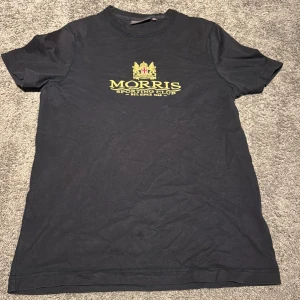 Mörk blå Morris t-shirt med tryck XS - Mörkblå  t-shirt från Morris Sporting Club i storlek XS. Klassisk rund hals och korta ärmar. Framsidan har ett gult tryck med Morris logga och emblem. Mjuk och skön bomull som passar perfekt till jeans eller shorts. Snygg och enkel design med stilren vibe.