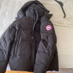 Canada Goose jacka - Säljer en svart dunjacka från Canada Goose med huva och klassisk logga på ärmen. Jackan har quiltad design, dragkedja och knappar framtill samt två stora fickor. Perfekt för kalla dagar och har en clean, stilren look. En knap har ramlat av, pris kan diskuteras
