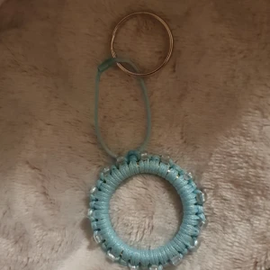 Handgjord blå nyckelring med pärlor - Unikt halsband med rund hängare i ljusblå snodd och transparenta pärlor runt hela cirkeln. Hängs i en enkel blå snodd och har en metallring upptill. Perfekt för dig som gillar personliga accessoarer med boho-vibe.
