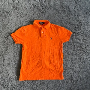 Ralph Lauren piké  - Snygg orange pikétröja från Polo Ralph Lauren med klassisk krage och två knappar framtill. Tröjan har korta ärmar och den ikoniska broderade loggan på bröstet. Tillverkad i mjuk bomull som andas, perfekt för en chill och sportig look.