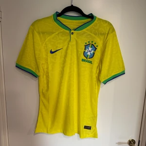 Brasilien Nike fotbollströja M - Gul Brasilien fotbollströja från Nike med blått Swoosh och CBF-emblem på bröstet. Tröjan har gröna och blå detaljer vid krage och ärmslut, samt ett subtilt leopardmönster i tyget. Tillverkad i lätt Dri-FIT material som andas, perfekt för match eller träning.