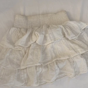 Vit volangkjol med resårmidja - Supersöt kort vit kjol med volanger och bred resår i midjan. Kjolen har ett diskret randigt mönster och är gjord i ett lätt, luftigt material som passar perfekt till varma dagar. Ger en romantisk och trendig vibe.