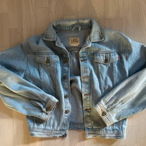 80's Vintage Jeansjacka  - Säljer denna vintage jeansjacka från tidigt 80-tal från Outdoor Jeanswear i storlek S. Jackan är ljusblå med autentisk fade, har två bröstfickor med knappar, snygga sömmar och bronsfärgade knappar framtill och vid ärmslut. Perfekt för dig som gillar retrovibe, truckerfits och vill ha en tidlös vibe.