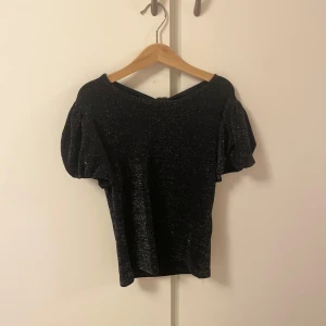 Glittrig svart topp med puffärm Lindex - Svart glittrig topp från Lindex med korta puffärmar och snygg knytning i ryggen. 