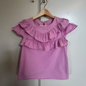 Ljusrosa blus/topp med volanger - Supersöt ljusrosa blus med dubbla volanger över axlar och bröst. Blusen har offshoulder-detaljer och rund halsringning med knapp i nacken.      Det finns ingen lapp, men jag tror att materialet är antingen linne eller bomull. Jag skulle säga att storleken är xs-s. 