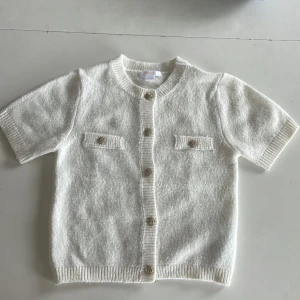 Vit stickad kofta från Gina Tricot - Supersöt vit stickad kofta från Gina Tricot med korta ärmar och rund hals. Koftan har snygga knappar framtill och två dekorativa fickor på bröstet. Perfekt att slänga över en topp för en chill och clean look.