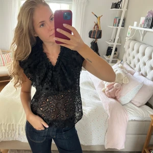 Svart spetsblus med volang - Säljer en svart blus med genombruten spets och volang vid axlarna. Blusen har v-ringad hals och är kortärmad med snygga detaljer i spetsen som ger en cool och feminin vibe. 