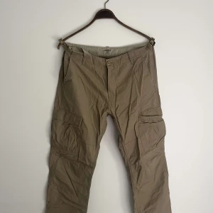 Beige cargopants från Carhartt WIP - Säljer ett par beige cargopants från Carhartt WIP med flera praktiska fickor på sidorna och bak. Byxorna har rak passform och är gjorda i slitstark bomull. Perfekt för dig som gillar streetwear och funktionella detaljer. nyskick, samt nypris 1199kr.