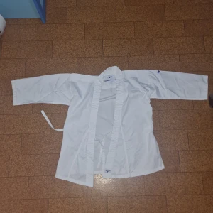 Vit karatekostym från Mizuno - Karatedräkt från Mizuno i helvitt med klassisk omlottjacka, raka byxor med snörning i midjan och tillhörande bälte. Kostymen är tillverkad i slitstark bomull och har Mizuno-logga i blått på jacka, byxa och bälte. Perfekt för träning eller tävling inom kampsport.