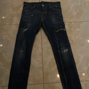 Mörkblå jeans från Dsquared2, strl 48 - Säljer ett par snygga mörkblå jeans från Dsquared2 i storlek 48. Jeansen har slitna detaljer och små hål för en cool, edgy look. Klassisk femficksmodell med raka ben och tydliga kontrastsömmar. Materialet är robust denim i bomull.