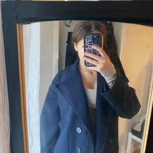 Mörkblå kappa från Design by Si - Snygg mörkblå kappa med klassisk krage och dubbelknäppning från Design by Si. Jackan har en loose passform och är långärmad, perfekt för kyliga dagar. Materialet känns som ullblandning och insidan är fodrad med svart tyg. Stilren och enkel att matcha. 