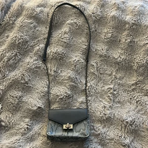 Grå axelväska med ormmönster från NYPD - Snygg axelväska från NYPD med kedjerem och lock i svart skinnimitation. Väskan har ett grått ormmönstrat material och coolt metallspänne framtill. På baksidan finns en rund metalllogga. Perfekt storlek för mobilen och det viktigaste.