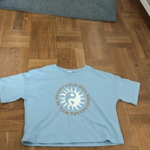 Ljusblå croppad t-shirt från Lindex - Snygg ljusblå croppad t-shirt från Lindex med korta ärmar. Framtill finns ett stort tryck med sol och yin-yang-symbol samt texten 'Sun will shine, stars will be bright'. Perfekt för dig som gillar chill och unik stil.