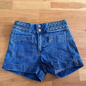 Blå jeansshorts från Never Denim - Snygga blå jeansshorts från Never Denim med flätad detalj i midjan och dubbla knappar framtill. Shortsen har stora fickor både fram och bak, och är tillverkade av bomull och polyester med lite stretch för skön passform.