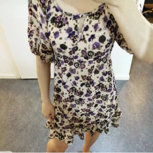 Blommig kortklänning med volang - Söt kortklänning med beige botten och blommigt mönster i lila och mörkbrunt. Klänningen har puffiga kortärmar, rund hals med liten öppning och knyt, samt volangkant längst ner. 