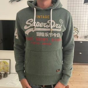 Grön Superdry hoodie med tryck - Snygg grön hoodie från Superdry med stort vintage-tryck i vitt, rött och gult på bröstet och siffran 54 på ärmen. Tröjan har huva med vita snören med gulddetaljer och en praktisk magficka. Perfekt för chill dagar och streetstyle.