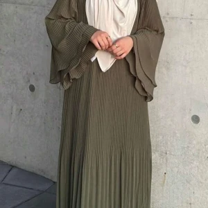 Plisserad långklänning i olivgrönt - Säljer en olivgrön långklänning med plisserat tyg och vida, vågiga ärmar. Klänningen har en elegant och flowy siluett som ger en chill vibe. Perfekt för dig som gillar stilrena detaljer och vill ha något som sticker ut med sin coola struktur. Aldrig använd. 
