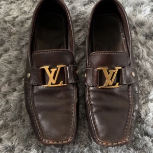Bruna Louis Vuitton loafers med LV-detalj - Snygga bruna loafers från Louis Vuitton i skinn med ikonisk LV-logga i guld på ovansidan. Skorna har fyrkantig tå, dekorativa sömmar och en platt sula med rutmönster undertill. Perfekta för dig som gillar lyxiga och klassiska detaljer.