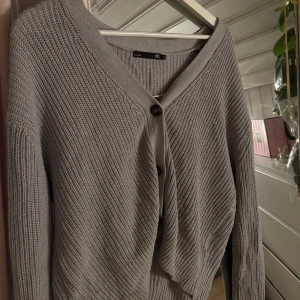 Grå stickad kofta från 157 - Säljer en grå stickad kofta från 157 med v-ringad hals och tre knappar framtill. Koftan har långärmade ärmar och ett grovt ribbat mönster. Perfekt att slänga över en t-shirt eller linne när det blir lite kyligt.