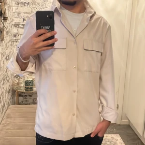 Beige overshirt med fickor - Snygg beige overshirt med klassisk krage och två stora bröstfickor med lock. Skjortan har knappar framtill och långa ärmar som kan rullas upp för en avslappnad look. Perfekt att bära över en t-shirt för en cool och enkel stil.