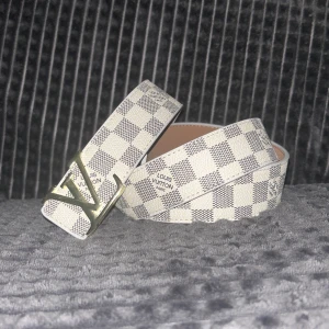 Rutig Louis Vuitton Vit 115cm - Säljer ett snyggt Louis Vuitton bälte med klassiskt rutigt mönster i vitt och grått. Bältet har en stilren LV-spänne i metall och är tillverkat i skinn med ljus insida. Perfekt för att lyfta din outfit med en exklusiv touch.