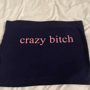 Svart kortkjol med rosa text - Svart kortkjol i mjukt bomullsmaterial med rak passform. Framsidan har ett tryck i ljusrosa med texten 'crazy bitch' som ger en edgy vibe. Perfekt för dig som vill sticka ut och visa attityd. Enkel design utan extra detaljer.