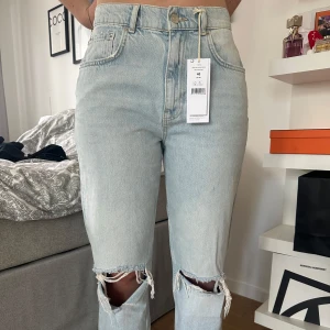 90s jeans ljusblå strl 40 - Säljer ett par 90-talsinspirerade jeans i ljusblå tvätt med raka ben och slitna hål vid knäna. Jeansen har klassisk femficksdesign, knapp och dragkedja framtill samt bälteshällor. Perfekt för dig som gillar retrovibe och relaxed fit.