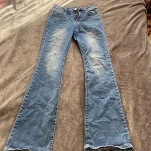 Blå bootcut jeans med detaljer - Säljer ett par blå bootcut jeans med snygga slitningar framtill och bakfickor med dekorativa knappar och mönstrad söm. Jeansen har klassisk femficksdesign och är tillverkade i mjukt denim. Perfekt för dig som gillar en retro vibe och vill sticka ut lite extra.