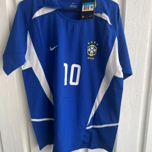 Blå Brasil Rivaldo fotbollströja Nike - Säljer en blå Brasilien fotbollströja från Nike med Rivaldo och nummer 10 på ryggen. Tröjan har vita detaljer och korta ärmar, samt Brasil-logga och Nike-swoosh på bröstet. Materialet är lätt och andas, perfekt för match eller träning.