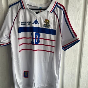 Frankrike Zidane VM 98 Adidas tröja - Säljer en ikonisk vit fotbollströja från Frankrikes VM 1998 med blå och röda detaljer, nummer 10 och Zidane på ryggen. Adidas-logga, broderat VM-märke och klassiska ränder över bröstet. Skön polyester med ribbade muddar och krage. Perfekt för dig som älskar retro och fotbollshistoria.