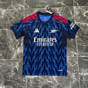 Arsenal blå zigzag fotbollströja Adidas - Snygg Arsenal fotbollströja från Adidas i mörkblått med elektriskt blått zigzag-mönster. Tröjan har korta ärmar och röda ränder på axlarna. Materialet är lätt och andas, perfekt för träning eller match. Emirates Fly Better-logga på bröstet och klubbmärke i vitt.