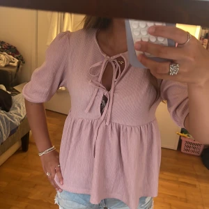 Ljusrosa blus med knyt och puffärm - Supersöt ljusrosa blus med knyt framtill och lätt puffade ärmar. Modellen är lite lös och har en flowy passform med rynk under bysten. Materialet är ribbat och toppen har en feminin, romantisk vibe. Perfekt till jeansshorts eller kjol för en chill look.