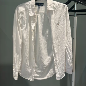 Vit skjorta från Ralph Lauren - Klassisk vit skjorta från Ralph Lauren med broderad logga på bröstet. Skjortan har lång ärm, knappar framtill och är tillverkad i ett lätt bomullstyg. Perfekt för en clean och stilren look.