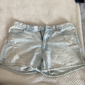 Ljusblå jeansshorts med pärlor Gina Tricot - Snygga ljusblå jeansshorts från Gina Tricot med pärldetaljer och slitningar framtill. Klassisk femficksmodell med knapp och dragkedja. Perfekta för sommaren och ger en cool vibe till din outfit.