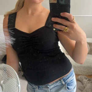 Svart ärmlös topp med spetsdetaljer - Säljer en svart ärmlös topp med breda axelband och snygga spetsdetaljer. Toppen har en lätt rynkning vid bysten och sitter figurnära. Perfekt att styla med jeans eller kjol för en trendig look.