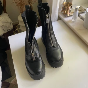 Chunky svarta boots med dragkedja - Säljer ett par chunky svarta boots med grov sula och dragkedja framtill. Skorna har ribbade detaljer på sidorna och är tillverkade i skinn och syntet. 