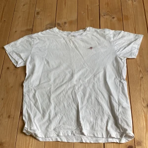 Vit t-shirt från GANT med broderi - Vit t-shirt från GANT i mjuk bomull med klassisk passform och rund hals. Liten broderad logga på bröstet ger en clean och stilren look. Perfekt till jeans eller shorts för en enkel och fräsch outfit.