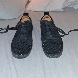 Svarta sneakers med nitar och röd sula - Unika svarta sneakers i mocka med coola svarta nitar på tån och klassisk röd sula undertill. Skorna har snörning och en låg profil, perfekt för dig som vill sticka ut. Insidan är ljusbeige och materialet känns lyxigt och robust. Storlek 44 passar 43