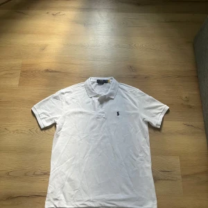 Vit pikétröja från Ralph Lauren - Klassisk vit pikétröja från Polo Ralph Lauren i custom slim fit. Tröjan har korta ärmar, krage och knäppning framtill samt den ikoniska mörkblå broderade loggan på bröstet. Perfekt för en clean och stilren look. Ny skick, sitter som M men funkar även som L. Inga defekter.
