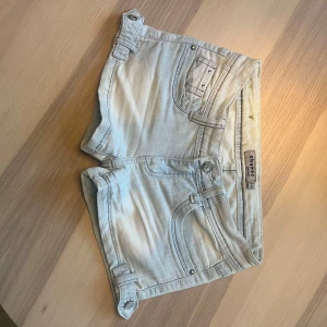 Ljusblåa jeansshorts från Cherri - Snygga ljusblå jeansshorts från Cherri med coola detaljer som nitar på bakfickorna och dekorativa knappar på sidorna. Klassisk femficksmodell med låg midja och slitna effekter. Perfekta för sommardagar när du vill ha en chill och trendig look. Många intresserade så var snabba! Kan gå ner i pris vid snabb affär!
