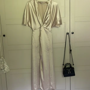 Champagnefärgad långklänning i satin - Elegant långklänning i skimrande champagnefärgad satin med djup v-ringning och korta, vida ärmar. Klänningen har en snygg twist-detalj framtill och ett mjukt, följsamt fall. Perfekt för dig som vill sticka ut med en lyxig och stilren look. ALDIRG använd!