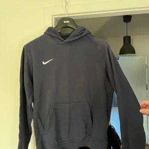 Mörkblå Nike hoodie med huva - Säljer en mörkblå hoodie från Nike i storlek L. Tröjan har klassisk huva, stor magficka och Nike-logga på bröstet. Perfekt för chill dagar eller när du vill ha en sportig look. Mjuk insida och långärmad modell.