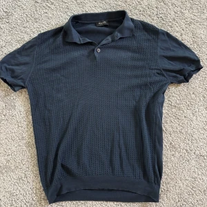 Mörkblå pikétröja från Sand Copenhagen - Snygg mörkblå pikétröja från Sand Copenhagen med klassisk krage och knappslå. Framsidan har ett diskret strukturerat mönster som ger en modern touch. Tröjan är kortärmad och tillverkad i mjuk bomull, perfekt för en stilren och avslappnad look.