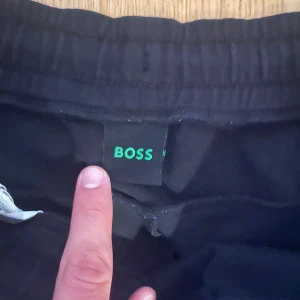 Svarta Boss shorts med snörning - Svarta shorts från Boss med broderad logga i vitt på benet. De har elastisk midja med snörning och sidofickor. Perfekta för en chill och sportig stil. Materialet känns mjukt och bekvämt, troligen bomull eller polyester.
