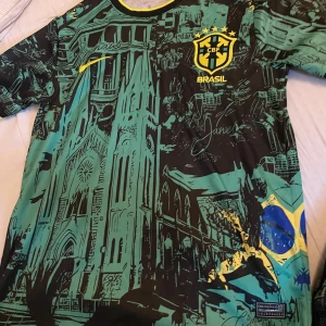 Nike Brasil CBF Dri-FIT fotbollströja - Unik Brasilien CBF fotbollströja från Nike med Dri-FIT-material. Tröjan har korta ärmar, svart och grön bas med gula detaljer och ett grafiskt tryck av brasilianska landmärken samt flaggan. Rund hals med svart och gul kant. Perfekt för dig som vill sticka ut på planen.