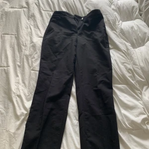 Svarta kostymbyxor från H&M - Svarta kostymbyxor från H&M i klassisk rak modell. Byxorna har midja med dragkedja och knapp, och är tillverkade i ett mjukt material som känns bekvämt hela dagen. Perfekta för en clean och stilren look.